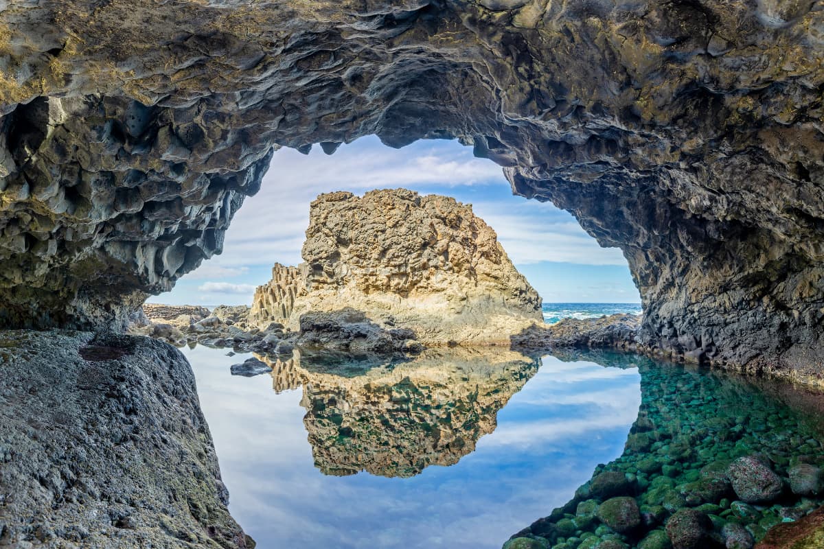 Esperienze 1 Vista dall'interno di una grotta marina rocciosa sulla Costa Occidentale Indomita di El Hierro, con lo sguardo rivolto a una grande formazione rocciosa che si riflette nell'acqua calma della marea, con il cielo azzurro e le nuvole sparse visibili all'esterno.
