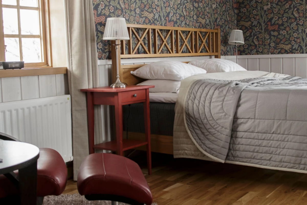 A neatly made bed with a gray quilt, red side table, bedside lamps, and patterned wallpaper creates a cozy bedroom with wood floors and natural light, capturing the inviting charm of Fryksås Hotell.
