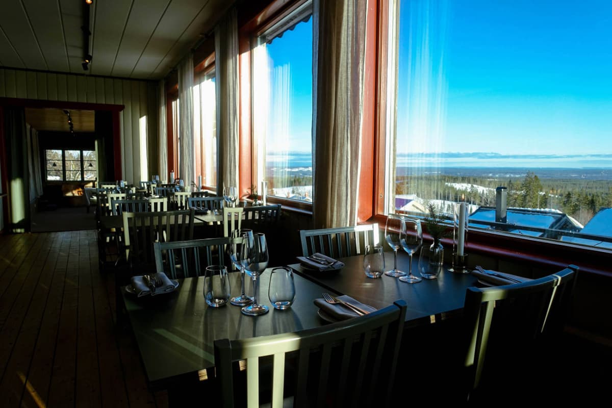 Restaurant dining area at Fryksås Hotell with set tables and large windows overlooking a scenic, sunlit landscape and clear blue sky.