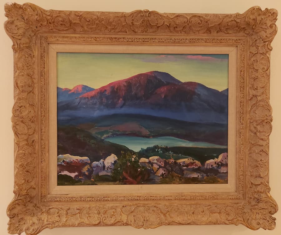 Belle come un quadro: cinque destinazioni europee che ispirano gli artisti 1 Un dipinto di paesaggio incorniciato che raffigura delle montagne con un lago in primo piano, sotto un cielo dalle sfumature blu e rosa.