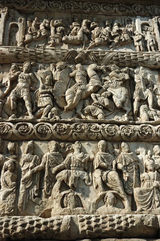 Belle come un quadro: cinque destinazioni europee che ispirano gli artisti 12 Rilievo in pietra dettagliato raffigurante numerose figure in abiti dell’antica Roma, impegnate in varie scene di raduno e combattimento, scolpito su una grande struttura architettonica.