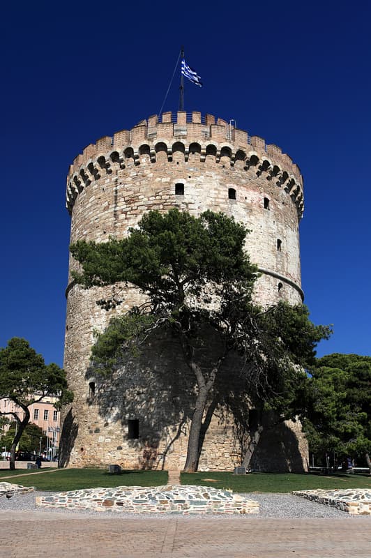 Belle come un quadro: cinque destinazioni europee che ispirano gli artisti 10 Una torre circolare in pietra, con merli e una bandiera greca in cima, si erge dietro un albero sotto un cielo azzurro e limpido.