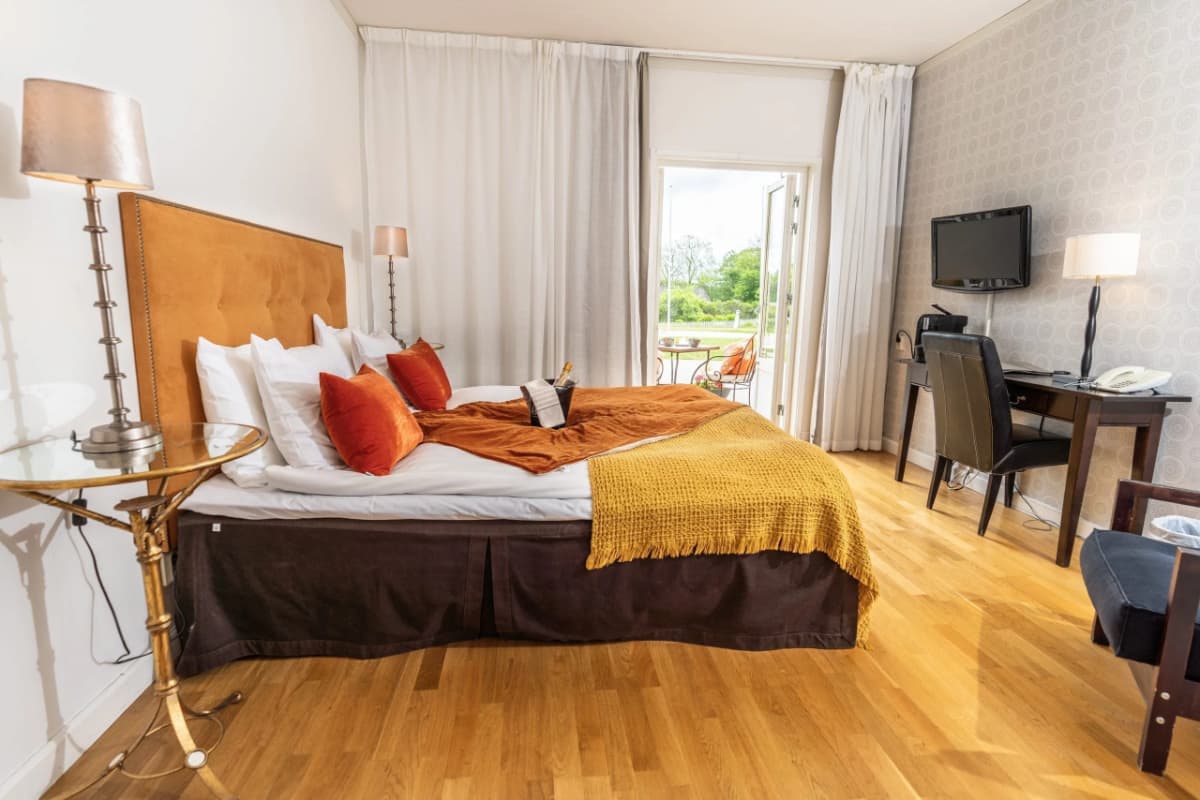 Moderna habitación del Körunda Hotell con cama doble, almohadas naranjas y blancas, una manta mostaza, escritorio y silla, TV y vistas panorámicas de la vegetación a través de puertas de cristal abiertas.