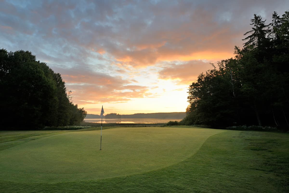 Green de golf con un palo de bandera al atardecer, rodeado de árboles y con vistas a un lago lejano bajo un cielo parcialmente nublado en Körunda Hotell.