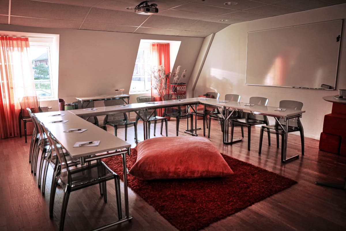 La moderna aula del Körunda Hotell cuenta con pupitres en forma de U, sillas blancas, un gran cojín rojo sobre una alfombra roja y una pizarra blanca en la pared. En cada pupitre hay blocs de notas y bolígrafos.