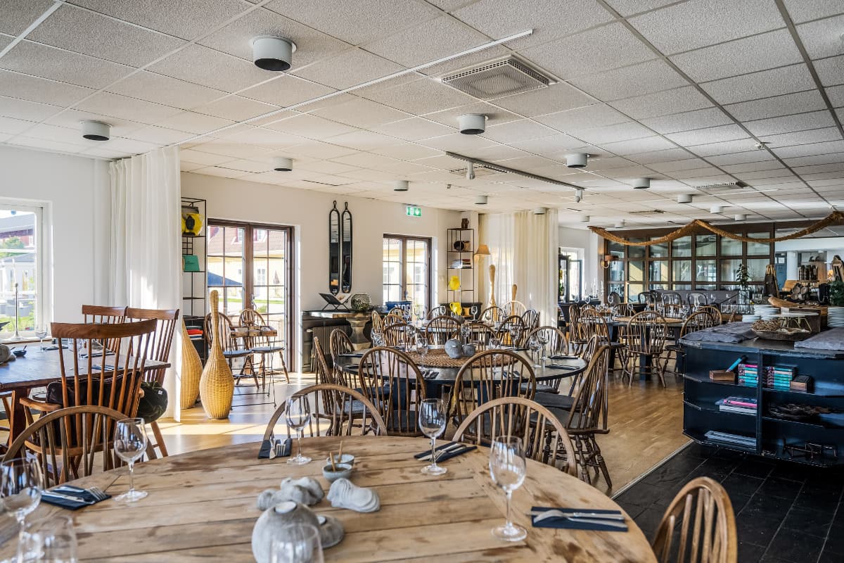 Amplia zona de comedor del restaurante Körunda Hotell con mesas y sillas de madera, cuidadosamente dispuestas con cristalería y servilletas, y luz natural que entra por grandes ventanales.