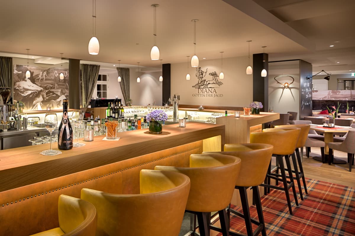 Modern bar with brown leather stools, wooden counter, pendant lights, and glassware. Tables and chairs are arranged in the background with decorative wall art and floral centerpieces.