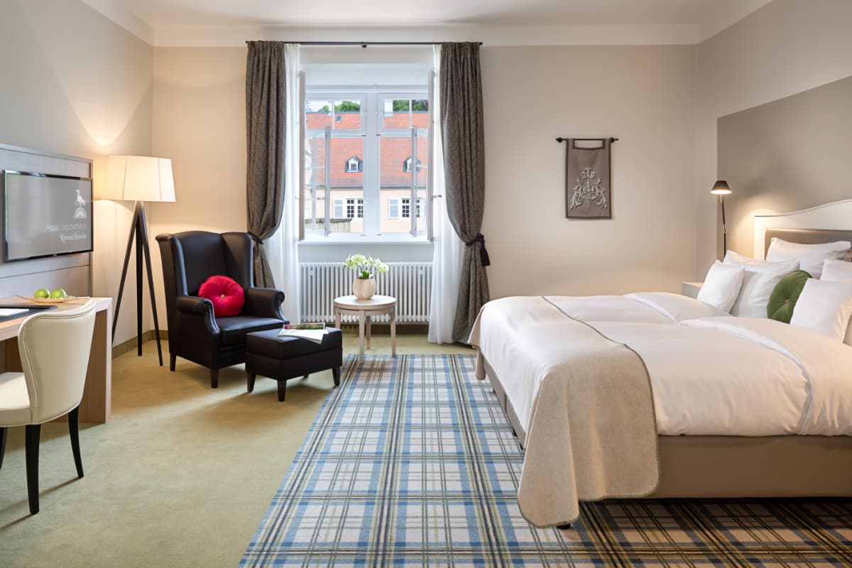 Modern hotel room with a double bed, armchair, desk, TV, patterned carpet, and a window with gray curtains overlooking rooftops.