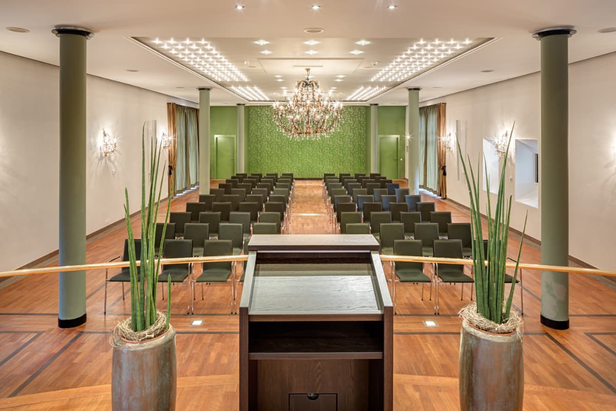 A modern conference room with rows of black chairs, a podium, green accent wall, chandelier, wood floors, and tall potted plants.