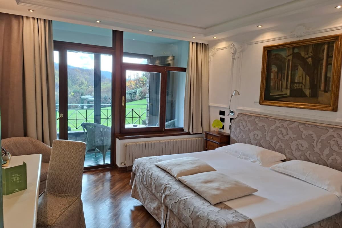 Hotel room with a double bed, two chairs, a desk, large window, and balcony overlooking a grassy area; a painting hangs above the headboard.
