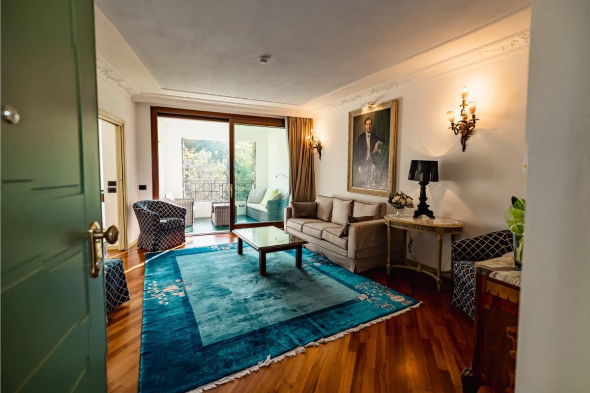 A living room with a blue rug, sofa, chairs, coffee table, wall art, and glass doors leading to a balcony with outdoor seating and greenery visible outside.