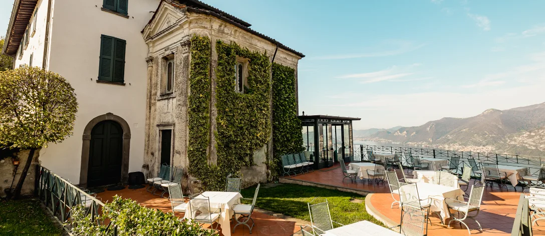 Outdoor terrace with white tables and chairs beside an old ivy-covered building, overlooking distant mountains and a lake under a clear sky.