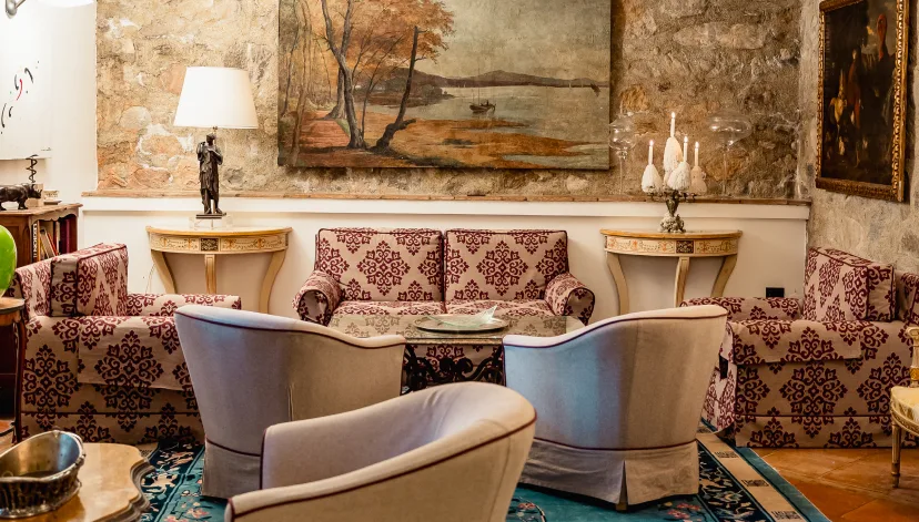 A cozy living room with patterned sofas, two armchairs, a large painting on a stone wall, two side tables, and a table lamp.