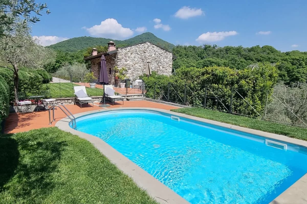 A rectangular outdoor pool with metal steps, surrounded by lawn, sun loungers, and a stone house with hills and trees in the background under a blue sky.