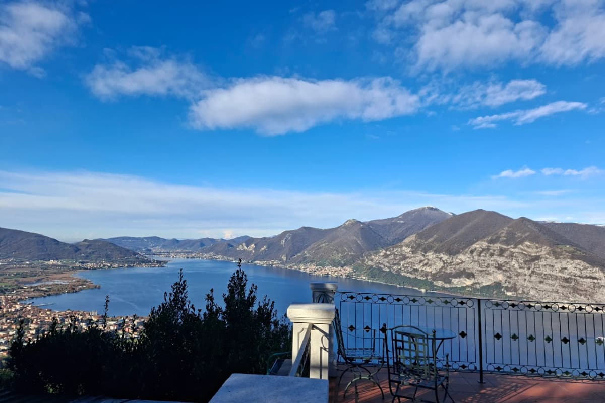 Eine Terrasse mit Tisch und Stühlen bietet unter einem teilweise bewölkten blauen Himmel einen Blick auf einen von Bergen umgebenen See und bietet den Gästen des 5-Sterne-Hotels „Iseo“, Relais I Due Roccoli, eine atemberaubende und ruhige Kulisse.