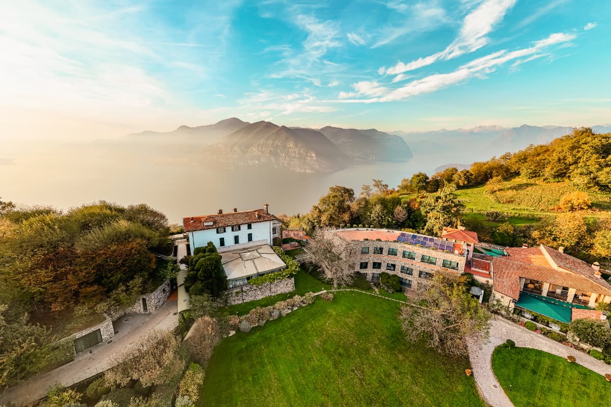Luftaufnahme des „Relais I Due Roccoli“, eines 5-Sterne-Hotels in Iseo, bestehend aus einem Villenkomplex mit Gärten und Blick auf einen großen See sowie die fernen Berge unter blauem Himmel.