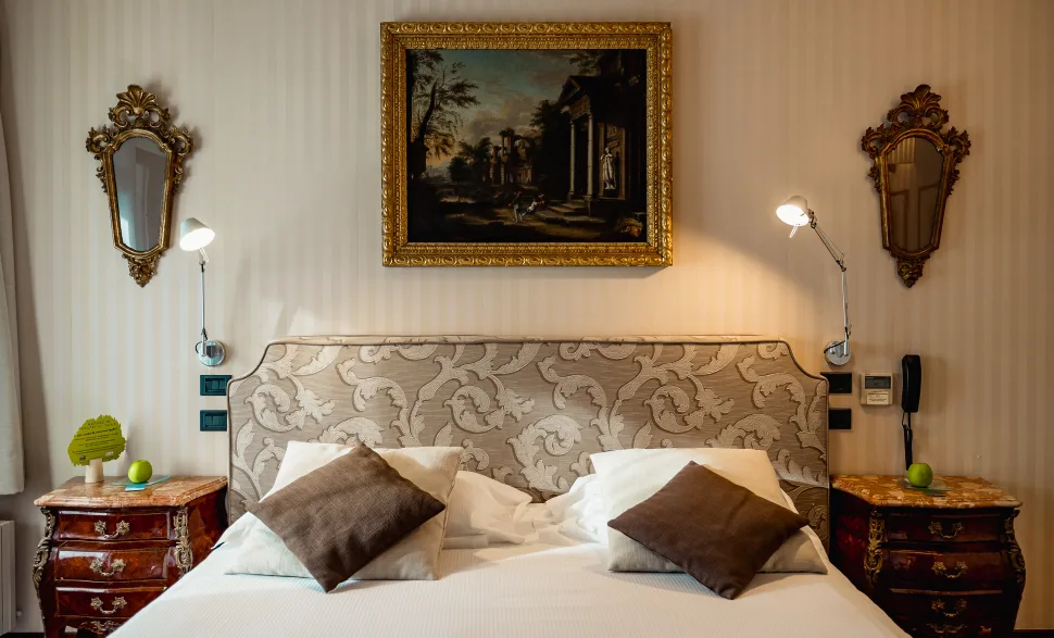 Ein ordentlich gemachtes Bett mit zwei braunen und zwei weißen Kissen, flankiert von zwei Nachttischen mit Lampen, dekorativen Spiegeln und einem Gemälde über dem Kopfteil – diese elegante Kulisse spiegelt das raffinierte Ambiente des Relais I Due Roccoli wider, eines renommierten 5-Sterne-Hotels in Iseo.