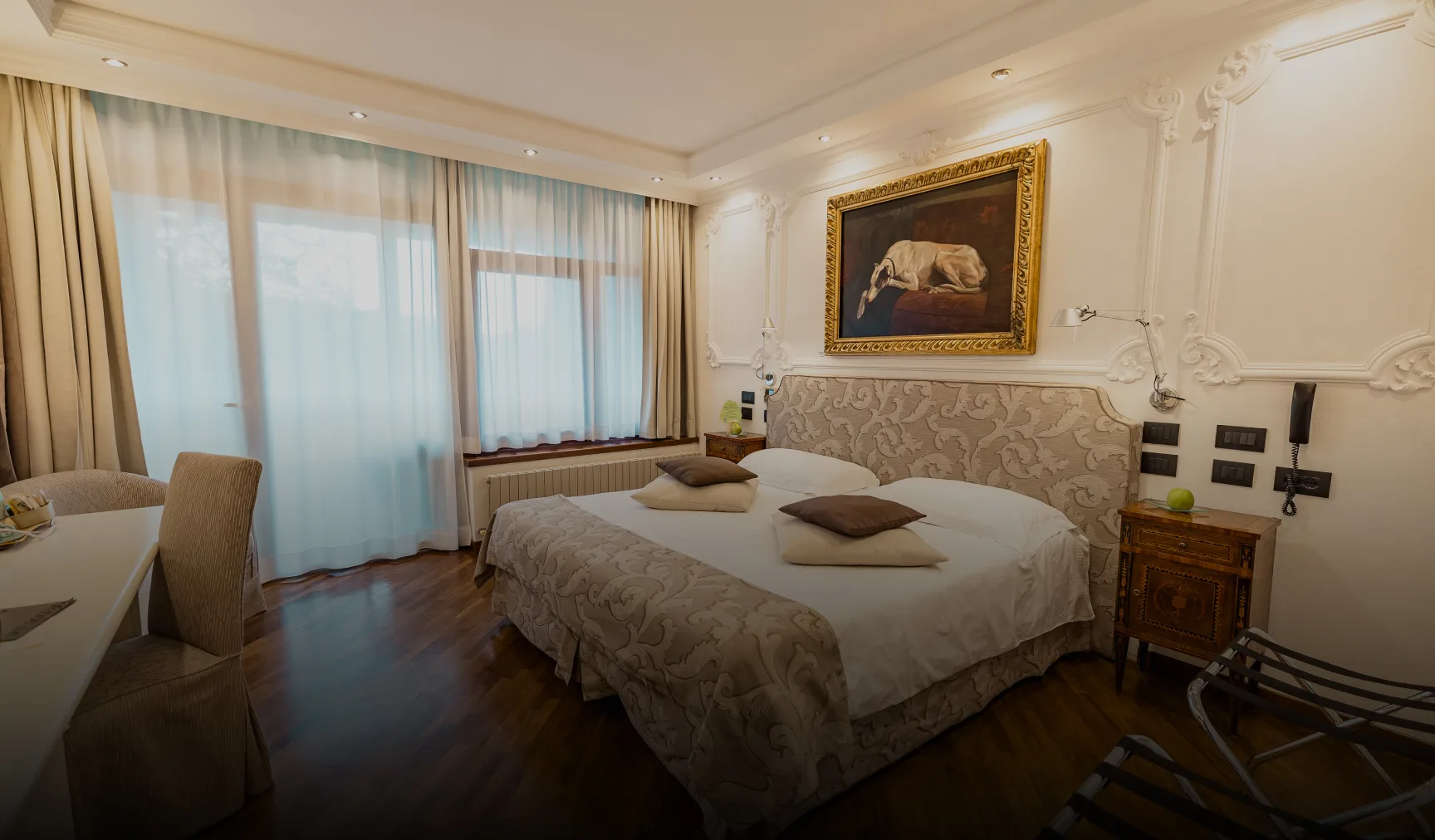 Ein Hotelzimmer mit einem Doppelbett, zwei Stühlen, einem Fenster mit Vorhängen, einem Schreibtisch und einem großen Gemälde, das einen liegenden Hund zeigt, über dem Bett – dieser einladende Raum im Relais I Due Roccoli verkörpert die raffinierte Eleganz, die man von einem 5-Sterne-Hotel in Iseo erwartet.