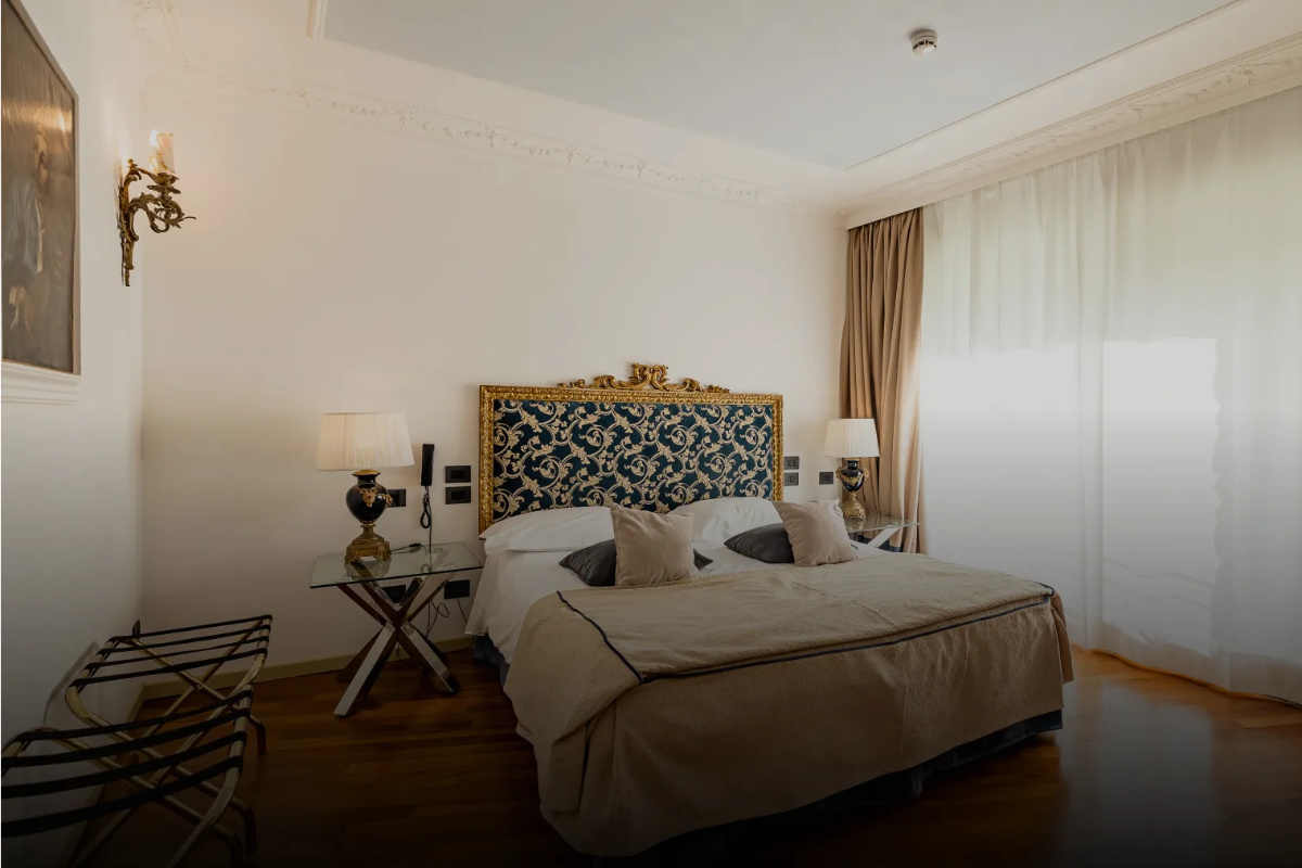 Ein Hotelzimmer im Relais I Due Roccoli, einem renommierten 5-Sterne-Hotel in Iseo, verfügt über ein Doppelbett mit einem kunstvoll verzierten Kopfteil, zwei Nachttische mit eleganten Lampen, einen Glastisch, einen Kofferabstellplatz sowie raumhohe Vorhänge, die viel Tageslicht hereinlassen.