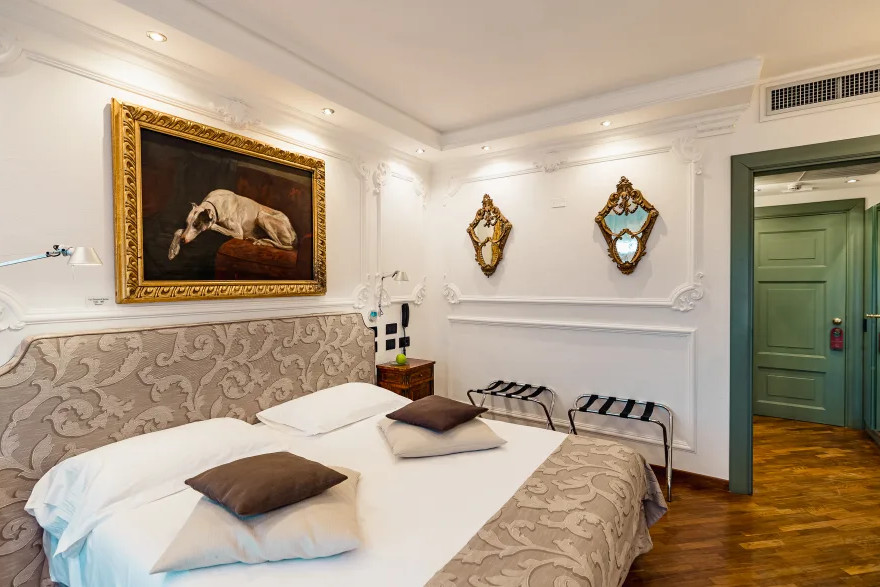In diesem hellen 5-Sterne-Zimmer des Hotels „Relais I Due Roccoli“ in Iseo erwarten Sie ein ordentlich gemachtes Doppelbett mit vier Kissen, kunstvolle, goldgerahmte Gemälde, zwei Spiegel und eine grüne Tür.