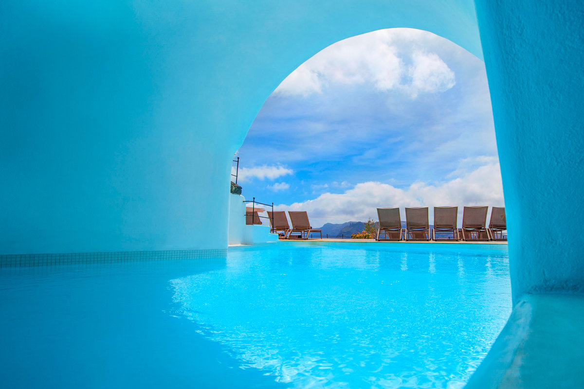 Una piscina coperta dalle pareti blu si affaccia su uno spazio all’aperto con sedie a sdraio, sotto un cielo parzialmente nuvoloso e con le montagne in lontananza.
