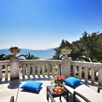 Cheque regalo 11 En el balcón junto al mar del Hotel Milenij hay dos tumbonas con cojines azules. Sobre la mesa, una botella de vino, copas y un cuenco de fruta. De fondo, se ven el océano y los árboles bajo un cielo azul despejado.