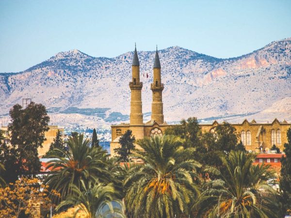 Eine steinerne Moschee mit zwei Minaretten steht zwischen Palmen, im Hintergrund erheben sich braune Berge unter einem klaren Himmel.