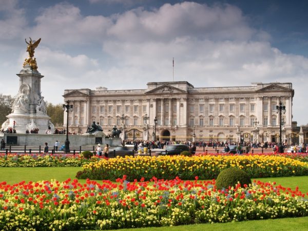El Palacio de Buckingham con el Victoria Memorial delante, rodeado de coloridos parterres y un cielo azul con nubes.