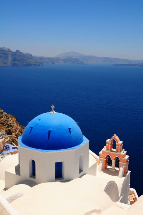 Un bâtiment blanc avec un dôme et un clocher bleus surplombe la mer d'un bleu profond sous un ciel clair sur la côte de l'île de Santorin, en Grèce.