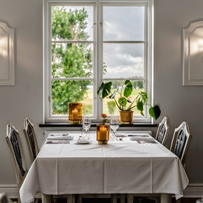 Cheque regalo 14 Una mesa de comedor para cuatro personas con mantel blanco y cristalería, situada junto a una ventana con plantas en macetas y dos apliques a cada lado.